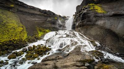 A waterfall cascades down rocky cliffs surrounded by green moss under a cloudy sky.