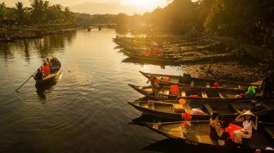 Several wooden boats with people wearing life jackets float on a river at sunset, with trees and buildings lining the water’s edge.