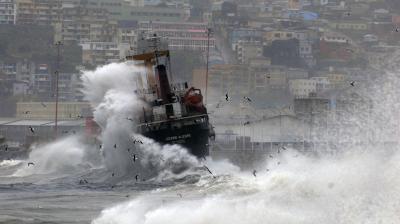 A ship being struck by large waves in a harbor, with a hillside city in the background.
