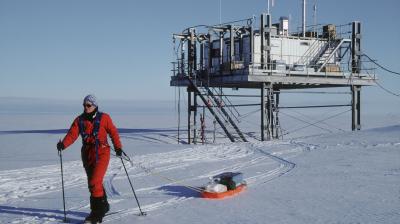 A person in a red suit cross-country skis across snow, pulling a loaded orange sled toward a raised research building.