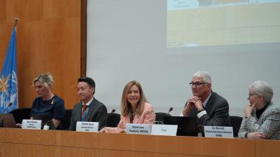 Five people sit at a conference table with nameplates, laptops, and microphones, in front of a large screen displaying a meeting presentation.