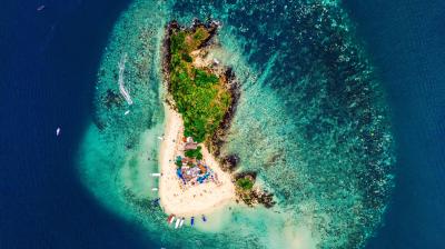 Aerial view of a small, lush island surrounded by a shallow coral reef and anchored boats.