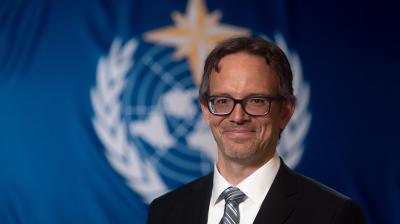 A man in a suit and glasses smiling in front of a blue United Nations flag.