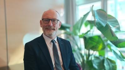 A smiling middle-aged man with a beard and glasses, wearing a dark suit and tie, sitting in an office with a large plant behind him.