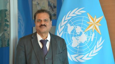 Man in a dark grey suit and patterned tie standing in front of a blue World Meteorological Organization flag.