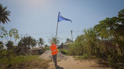 A person in an orange vest carries a tall pole with a blue flag on a dirt path, with trees and houses in the background under a clear sky.