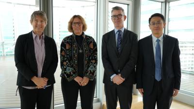 Four people in business attire standing in a row in a bright, modern room with large windows.