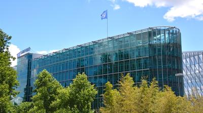 A multi-story office building with a glass facade and a blue flag flying from the roof, viewed behind trees.