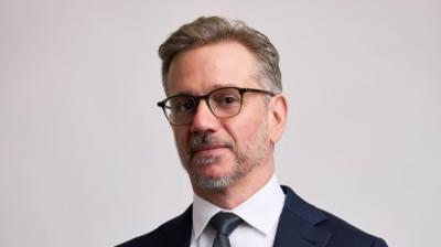 Man wearing glasses and a dark suit with a white shirt and tie stands against a plain light background, looking at the camera