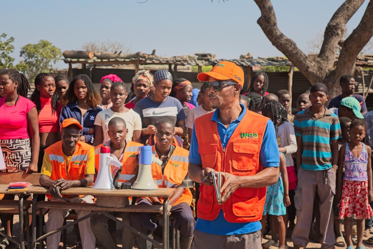 A group of people, including several in orange vests, stand outdoors near tables with megaphones. One person in an orange cap addresses the crowd.
