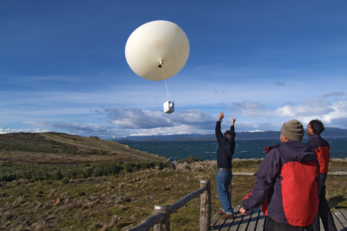Three people stand on a wooden platform as a large white weather balloon with attached equipment ascends into the sky over a grassy, hilly landscape.