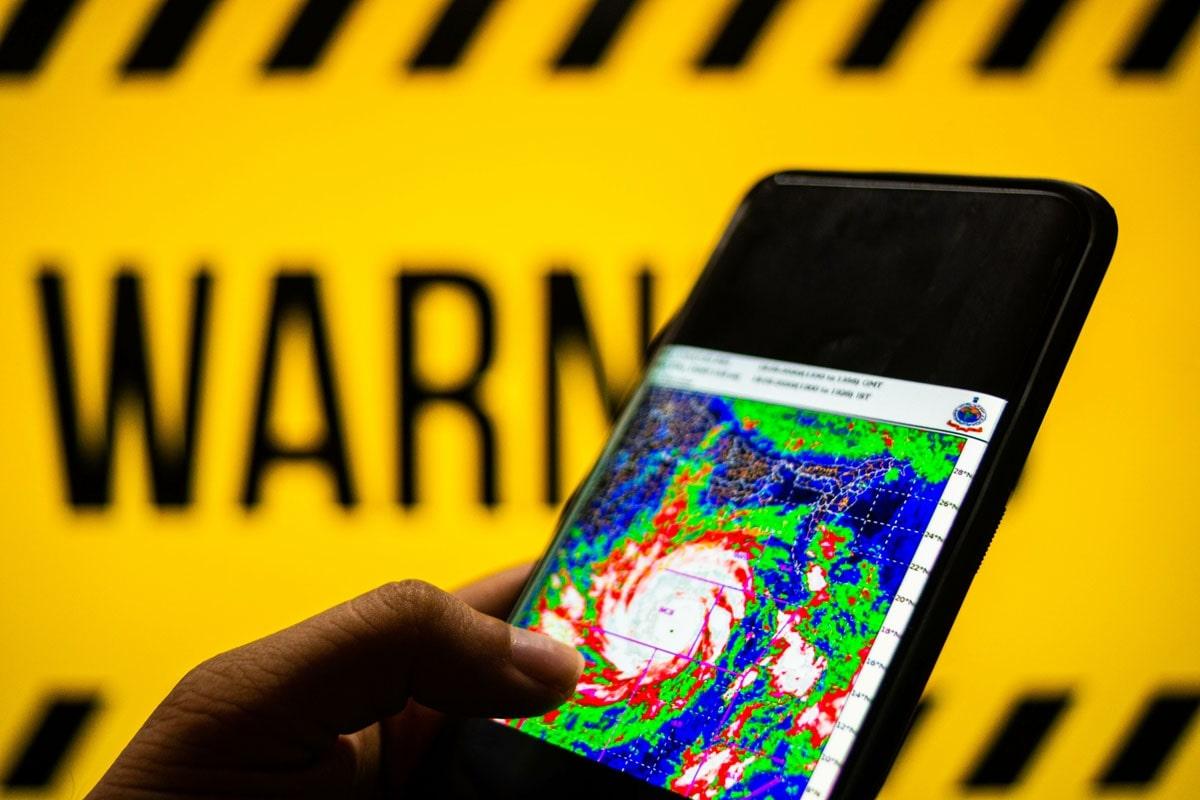 A hand holds a smartphone displaying a weather radar map in front of a yellow background with the word "WARNING" in bold black letters.
