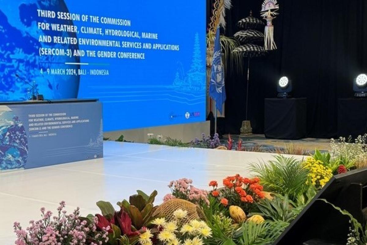 A conference stage with a large blue screen displaying information about the Third Session of the Commission for weather and climate in Bali, decorated with colorful flowers in the foreground.