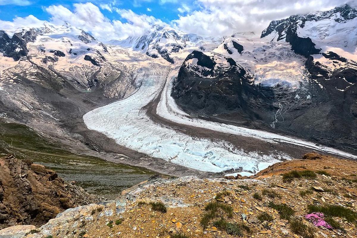 A wide glacier winds through a rocky mountain valley under a partly cloudy sky, with patches of snow on the surrounding peaks.