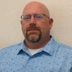 A bald man in a blue shirt standing next to a wall.