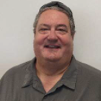 A man in a gray shirt smiling in front of a white wall.