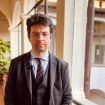 Alberto Pirni in a suit and tie standing in front of a window.