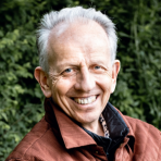 An older man smiling in front of bushes.