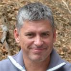 A man in a gray jacket smiling in front of a wooded area.