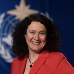 A woman in a red jacket smiling in front of a flag.