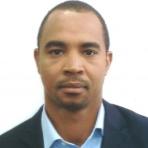 A man in a suit and blue shirt is standing in front of a white wall.