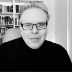 A black and white portrait of a bespectacled man with a slight smile, wearing a turtleneck, in front of a bookshelf.