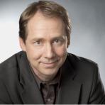 A man in a black jacket and shirt smiling at the camera with a grey background.