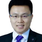 A man with short black hair, wearing glasses, a dark suit, a white shirt, and a purple tie, smiles at the camera against a plain white background.