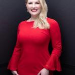 A woman with long blonde hair, in a red dress with bell sleeves, stands against a dark, plain background, smiling at the camera.