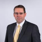 A man in a dark suit, light yellow tie, and glasses poses against a plain white background.