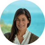 A woman with glasses and long hair wearing a white blouse and gray cardigan stands in front of a blue background.