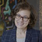 A person with curly hair and glasses smiles, wearing a checkered blazer. A colorful abstract background is visible.