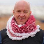 A person wearing a red and white keffiyeh around their neck, standing outdoors, smiling at the camera.