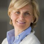 A woman with short blonde hair smiles, wearing a blue collared shirt and a white textured jacket.