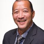 Man in a dark suit and patterned shirt smiling against a plain background.