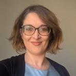 A person with shoulder-length hair, wearing glasses and a light blue top with a black cardigan, smiles at the camera.