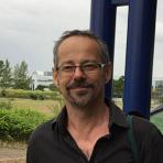 A man with glasses and short hair stands outdoors, wearing a black shirt. There is greenery and a large dark blue structure in the background.