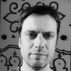 Man with short hair and a neutral expression standing in front of a patterned wall. Black and white photo.