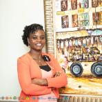 Smiling person in an orange blazer stands with arms crossed in front of a colorful painting depicting a market scene. Various organizational logos are visible at the bottom edge.