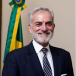 Smiling man in a suit and tie stands in front of a wall, with a partially visible flag in the background.