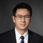 A man in a suit and tie, wearing glasses, looking at the camera with a neutral expression against a dark background.