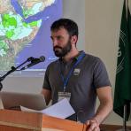 Man speaking at a podium with a microphone, laptop, and papers, in front of a world map projection. Green and white regions highlight areas on the map. Flag partially visible on the right.