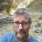 A person with glasses and a beard takes a selfie outdoors in a rocky, wooded area, wearing a blue shirt.