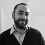 Man with a beard smiling, wearing a collared shirt and sweater, in a black and white photo.