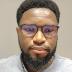 A man with short curly hair, a beard, and glasses is wearing a light grey shirt and looking at the camera against a plain background.