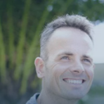 A man with short hair smiling outdoors, with a blurred background of green foliage and blue sky.
