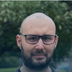 A person with glasses and a beard is outdoors with trees in the background.