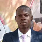 A man in a dark suit and light pink tie stands in front of a backdrop featuring a woman in graduation attire making a hand gesture.