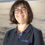 A woman with glasses and short brown hair smiles while wearing a navy polo shirt with the SWOT logo, standing in front of a backdrop showing clouds and space.
