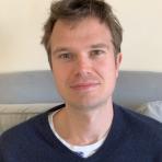 A man with short brown hair and blue eyes sits on a gray couch, wearing a navy blue sweater over a white shirt, facing the camera in a well-lit room.